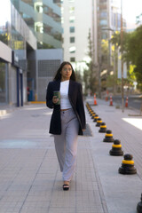 Close-up of young Hispanic businesswoman walking on city sidewalk with mug in hand, wearing blue blazer, white shirt, and heels