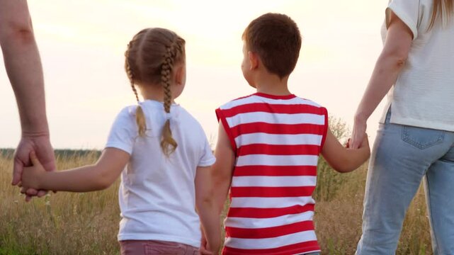 Happy little children walk joining hands with parents across summer meadow at sunset backside view. Loving children son and daughter discover nature with parents. Family and children walk in field