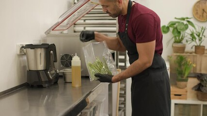 Chef prepares a plastic bag with olive oil and Asparagus for vacuum sealing in his professional kitchen. The setting features a clean workspace with fresh ingredients. Medium shot, side view.
