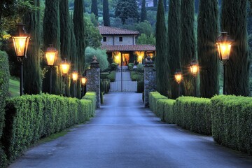 Fototapeta premium Elegant pathway illuminated by classic lamps, leading to a charming villa amidst cypress trees in a serene countryside setting at dusk, creating a magical atmosphere