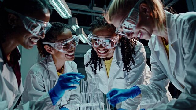 Grupo de estudiantes latinas con batas blancas y gafas protectoras sonr&iacute;en mientras colaboran en pruebas cient&iacute;ficas en un laboratorio educativo.
