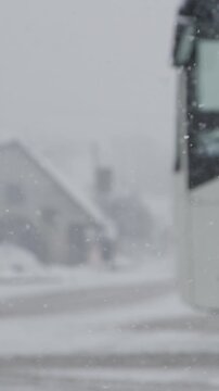 SLOW MOTION, DOF: White bus slows down at a bus stop during a snowy winter day. Bad weather gathers over the city streets and covers them in snow. Tourist coach stops due to bad weather conditions.