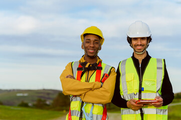 Two wind energy workers wearing safety vests and helmets are smiling while holding a digital tablet...