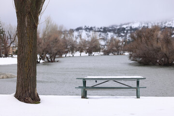 A horizontal presentation of a landscape image with a park bench, a tree, a lake and snow in the blurred background.