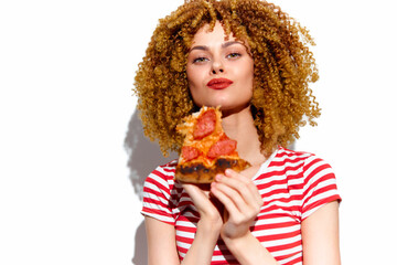 Young woman with curly hair smiling while holding a slice of pizza, dressed in a striped shirt, showcasing a joyful and casual lifestyle