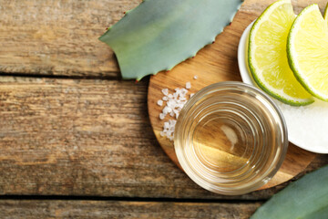 Tequila shot with lime slices, salt and agave leaves on wooden table, flat lay. Space for text