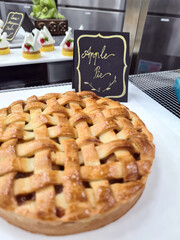 A freshly baked apple pie with a golden lattice crust sits proudly in a bakery, accompanied by an elegant handwritten sign
