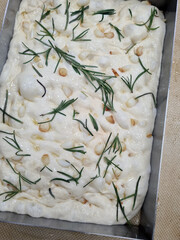 Freshly made focaccia bread topped with rosemary and pine nuts is ready for baking, showcasing the ingredients and preparation style