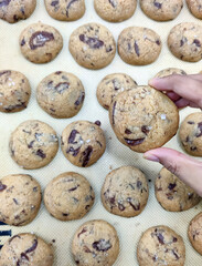 A hand holds a chocolate chip cookie filled with melted chocolate, with more cookies cooling on a tray nearby