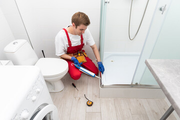 Young man in uniform repairs the shower door in the bathroom. A male repairman repairs the shower cabin.