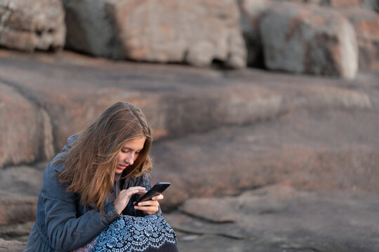 Girl huddled in jacket outdoors looking at smartphone - Powered by Adobe