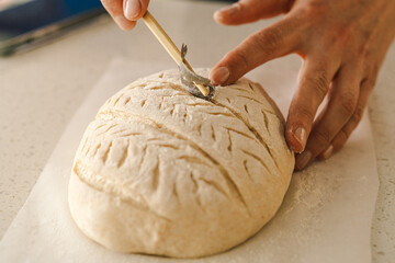 Woman hands skillfully score the surface of a round dough loaf, preparing sourdough bread for baking. The bright kitchen setting provides a warm atmosphere and fresh ingredients.