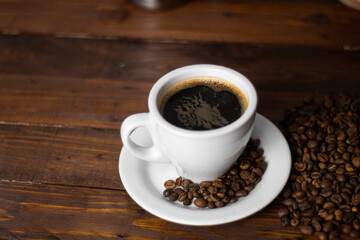 A glass of white mug with a white saucer served on a wooden background with coffee beans beside it. top view. Cup coffee beans wooden. Black coffee in white cup with coffee beans. copy space coffee