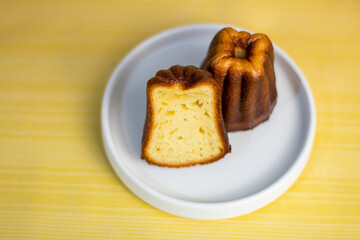 Close up of Canele, French pastry flavored with rum and vanilla, soft and tender custard center and dark, canele served with half cut, white plate, coffee, books, coffee beans and yellow background