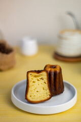 Close up of Canele, French pastry flavored with rum and vanilla, soft and tender custard center and dark, canele served with half cut, white plate, coffee, books, coffee beans and yellow background