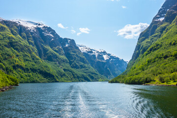 Snowcapped mountains lined with lush green trees and grass, inside of the fjord, N&aelig;r&oslash;yfjord, in Norway, Scandinavia, on a blue sky day with minimal clouds