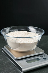 Electronic kitchen scale with bowl of flour on dark textured table against black background, closeup