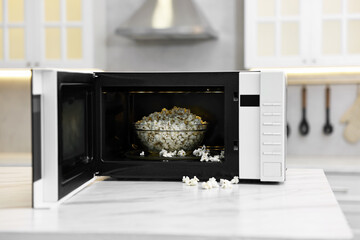 Open microwave oven with bowl of popcorn on white marble table in kitchen