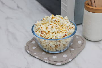 Tasty popcorn in bowl near microwave oven on white marble table, closeup