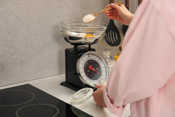 Woman adding flour into bowl on mechanical kitchen scale at white marble countertop indoors, closeup