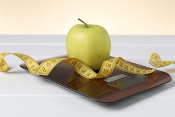 Electronic kitchen scale, measuring tape and apple on white wooden table against beige background,...