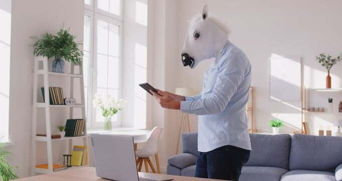 Male corporate worker in horse mask being half animal, using modern technologies in his work, holding tablet and touching screen, searching for information online, standing in home room