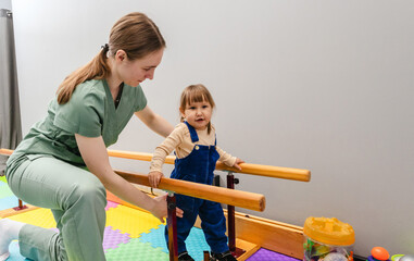 Little female child is engaging in physical therapy, assisted by a caregiver, using parallel bars to improve mobility. Handrails for restoring walking skills for children
