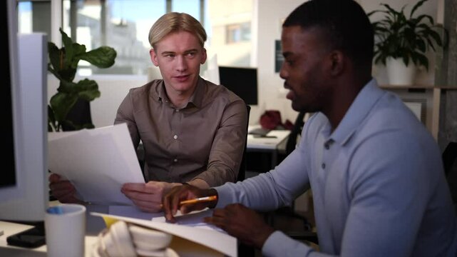 Two young male professionals, one caucasian and one african american, collaborate on a project, reviewing paperwork together at a shared desk in a contemporary office environment