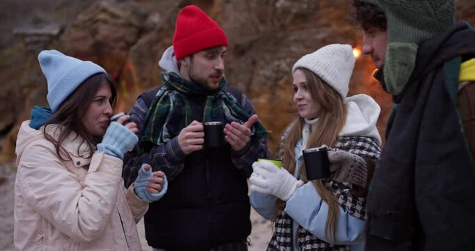 Happy friends in warm clothes drink tea from cups and chat outside the home