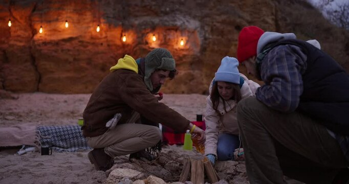 A guy pours lighter fluid on a fire during a break with his friends on a cozy beach