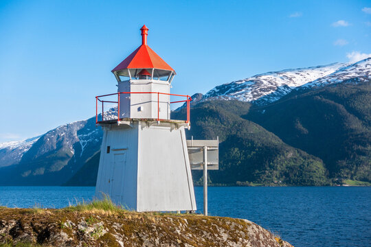 Close up of lighthouse on rocky hill along coast of vintage Norwegian town Leikanger along Norway's Sognefjord with blue skies small clouds and green snowcapped mountains in background