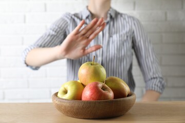 Woman refusing to eat apples at wooden table near white brick wall, selective focus. Food allergy...