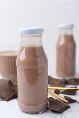 Tasty chocolate milk in glass bottles and pieces of chocolate on white tiled table, closeup