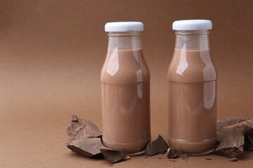 Tasty chocolate milk in glass bottles and pieces of chocolate on brown background, closeup. Space for text