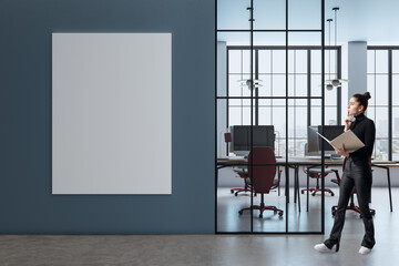 Modern office interior with blank vertical poster on dark blue wall and businesswoman holding...