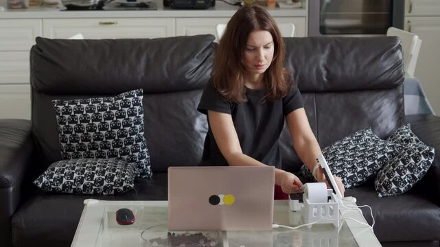 A woman using a laptop and label printer to prepare for her online business, working from the comfort of her living room with decorative pillows on the sofa.
