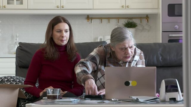A middle-aged woman patiently instructs her elderly mother on using a laptop and thermal label printer for their online store business, set in a cozy home environment.