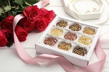 Flower shaped chocolate bonbons in box, red roses, pink ribbon and pearl jewelry on white wooden table, closeup