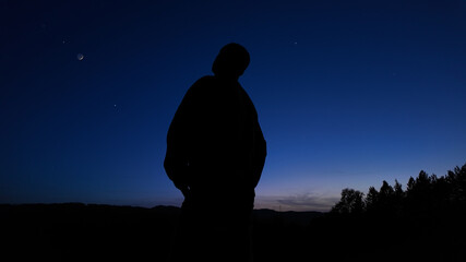 Silhouette of a man and landscape under the stars, planets, Moon and shooting-star trails.