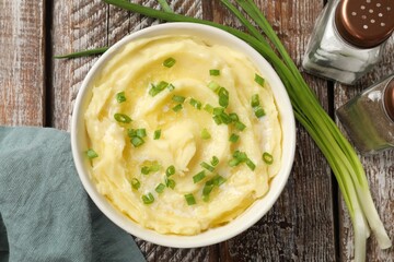 Tasty mashed potato with green onion in bowl and spices on wooden table, flat lay