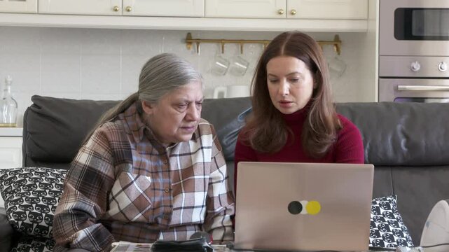 A daughter explains the nuances of online trading to her elderly mother on a laptop, fostering digital empowerment and strengthening their intergenerational bond in the familiar setting of their home.