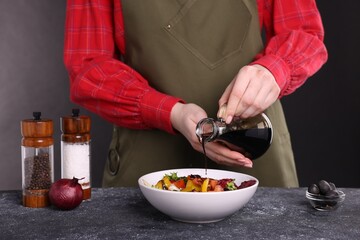 Woman pouring balsamic vinegar onto tasty salad at dark textured table, closeup
