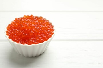 Tasty red caviar in bowl on white wooden table, closeup. Space for text