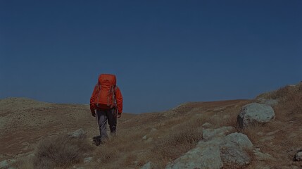 Fototapeta premium A hiker with a vibrant orange backpack traverses a dry, mountainous landscape. The hiker is seen from behind, walking away from the camera, through scrub-covered hills