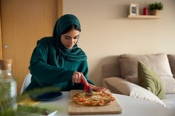 Muslim woman cutting pizza at home with pizza cutter