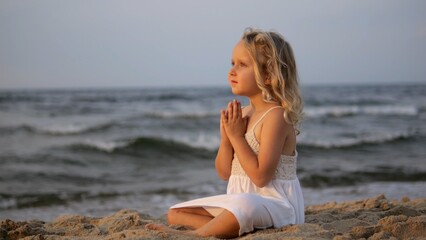 Blond girl wearing white dress sitting peacefully, meditating with hands together while facing ocean during golden sunset, embodying tranquil childhood innocence. concept of faith since childhood