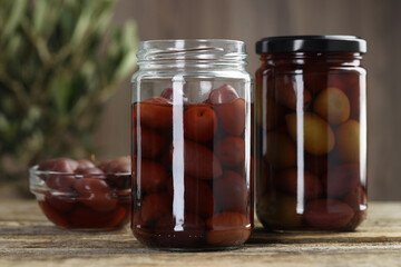 Pickled olives in glass jars and bowl on wooden table, closeup