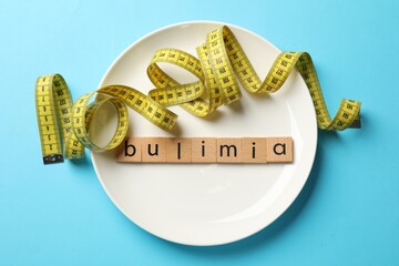 Plate with word Bulimia and measuring tape on light blue background, top view
