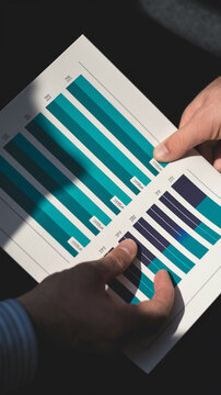 Close-up of hands reviewing teal and dark-grey bar chart on paper, showcasing business analysis and data review concept