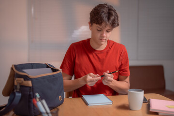 Fototapeta premium Teenager playing with a pen while sitting at a desk with notebooks, laptop, smartphone and a cup of coffee, preparing for exams or doing homework